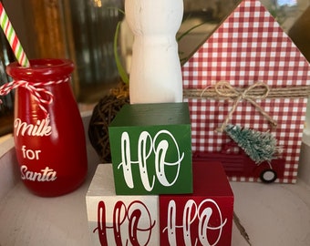 May include: A tiered tray decorated for Christmas with a red and white gingham house, a green block with white lettering that says "HO HO", and a red block with white lettering that says "HO HO".