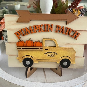 May include: A wooden sign with a white background and brown accents. The sign says "Pumpkin Patch" and features a yellow truck with a bed full of orange pumpkins. The truck has the words "Harvest Acres Farm" on the side.