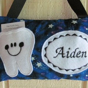May include: A blue tooth fairy pillow with a white tooth and a white circle with the name Aiden embroidered on it. The pillow has a pocket with a note that says "Dear Tooth Fairy, in this pocket you will find..."