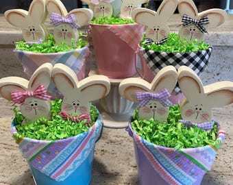 May include: Six wooden bunny figurines in small buckets with pastel colored fabric. The bunnies are painted white with pink and black accents. The buckets are filled with green shredded paper.