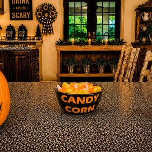 May include: A black ceramic bowl filled with yellow, orange, and white candy corn. The bowl features the text CANDY CORN printed in orange letters on the front. The bowl is positioned on a speckled countertop next to a carved orange pumpkin.