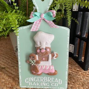 May include: A decorative, mint green cutting board with a gingerbread man chef and child design. The board features a pink and mint green bow and the text "GINGERBREAD BAKING CO" in white lettering.