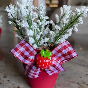May include: A small red pot with a white and red gingham bow and a red strawberry decoration. The pot is filled with white artificial flowers.