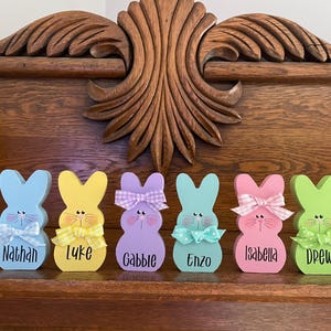 K&ouml;nnte beinhalten: Sechs farbenfrohe Holz-Osterhasenfiguren mit personalisierten Namen. Jeder Hase ist in einer anderen Pastellfarbe bemalt und hat eine Schleife. Die Namen sind in schwarzer Schrift geschrieben. Auf einer Holzoberfl&auml;che ausgestellt.