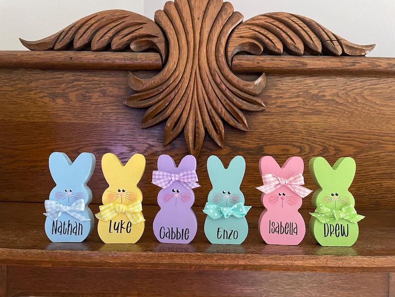 K&ouml;nnte beinhalten: Sechs farbenfrohe Holz-Osterhasenfiguren mit personalisierten Namen. Jeder Hase ist in einer anderen Pastellfarbe bemalt und hat eine Schleife. Die Namen sind in schwarzer Schrift geschrieben. Auf einer Holzoberfl&auml;che ausgestellt.