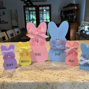 K&ouml;nnte beinhalten: Eine Sammlung von h&ouml;lzernen Osterhasen-Dekorationen in verschiedenen Pastellfarben. Jeder Hase hat eine Schleife und einen Namen, darunter "Mom", "Dad", "Roxy", "Melissa", "Maci" und "Jay", aufgeschrieben.