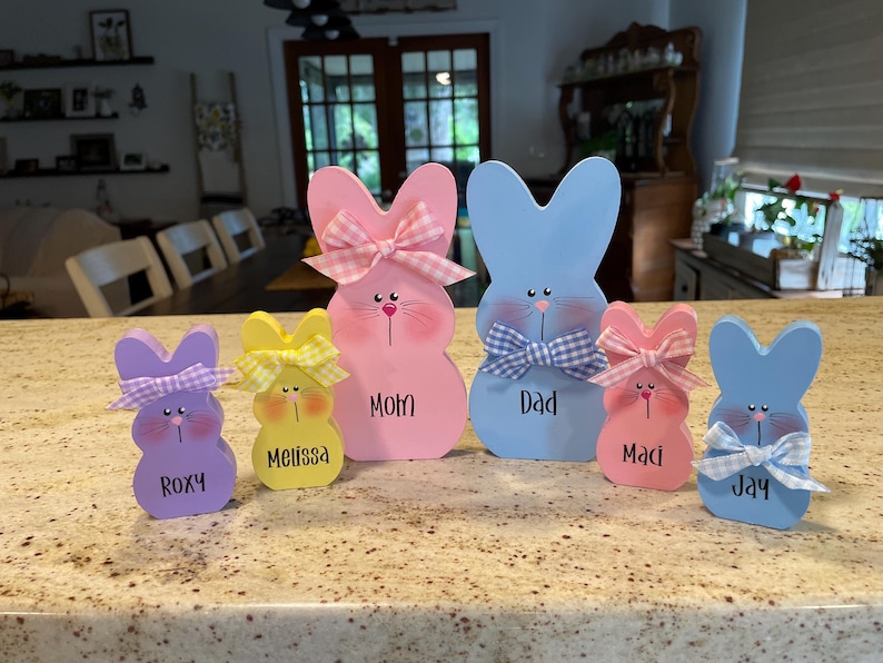 K&ouml;nnte beinhalten: Eine Sammlung von h&ouml;lzernen Osterhasen-Dekorationen in verschiedenen Pastellfarben. Jeder Hase hat eine Schleife und einen Namen, darunter "Mom", "Dad", "Roxy", "Melissa", "Maci" und "Jay", aufgeschrieben.