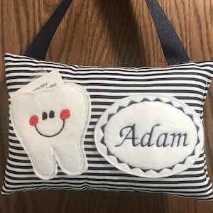 May include: A white tooth pillow with a smiling face and red cheeks. The pillow has a blue and white striped fabric and a white oval with the name "Adam" embroidered on it.