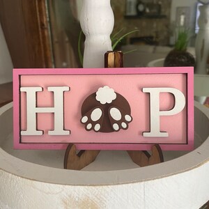 May include: A pink wooden sign with the word "HOP" in white letters. The "O" is replaced with a brown bunny butt with a white tail and white feet.