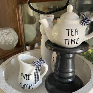 May include: White tiered tray with a black and white checkered bow, a white teapot with the text "TEA TIME", and a white creamer with the text "SWEET CREAM".
