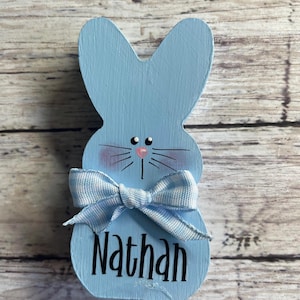 K&ouml;nnte beinhalten: Hellblaue Holz-Osterhasen-Dekoration mit Schleife und dem Namen "Nathan" in schwarzer Schrift. Der Hase hat rosa Wangen, schwarze Schnurrhaare und eine rosa Nase. Die Schleife ist hellblau und wei&szlig; kariert.