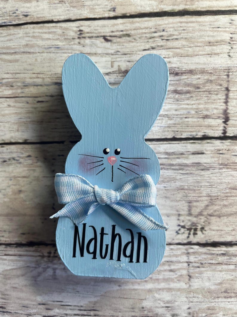 K&ouml;nnte beinhalten: Hellblaue Holz-Osterhasen-Dekoration mit Schleife und dem Namen "Nathan" in schwarzer Schrift. Der Hase hat rosa Wangen, schwarze Schnurrhaare und eine rosa Nase. Die Schleife ist hellblau und wei&szlig; kariert.