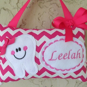 May include: A pink and white chevron tooth fairy pillow. The pillow has a smiling tooth applique with pink cheeks and a bow. The name "Leelah" is embroidered on a white oval. A pink ribbon is attached for hanging.