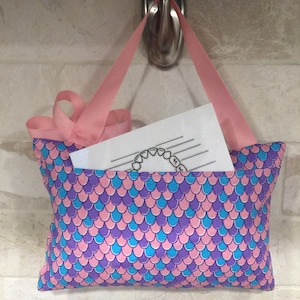 May include: A pink fabric bag with a mermaid scale pattern in pink, blue, and purple. The bag has pink ribbon handles and is hanging on a silver hook. A white piece of paper with a handwritten note is inside the bag.