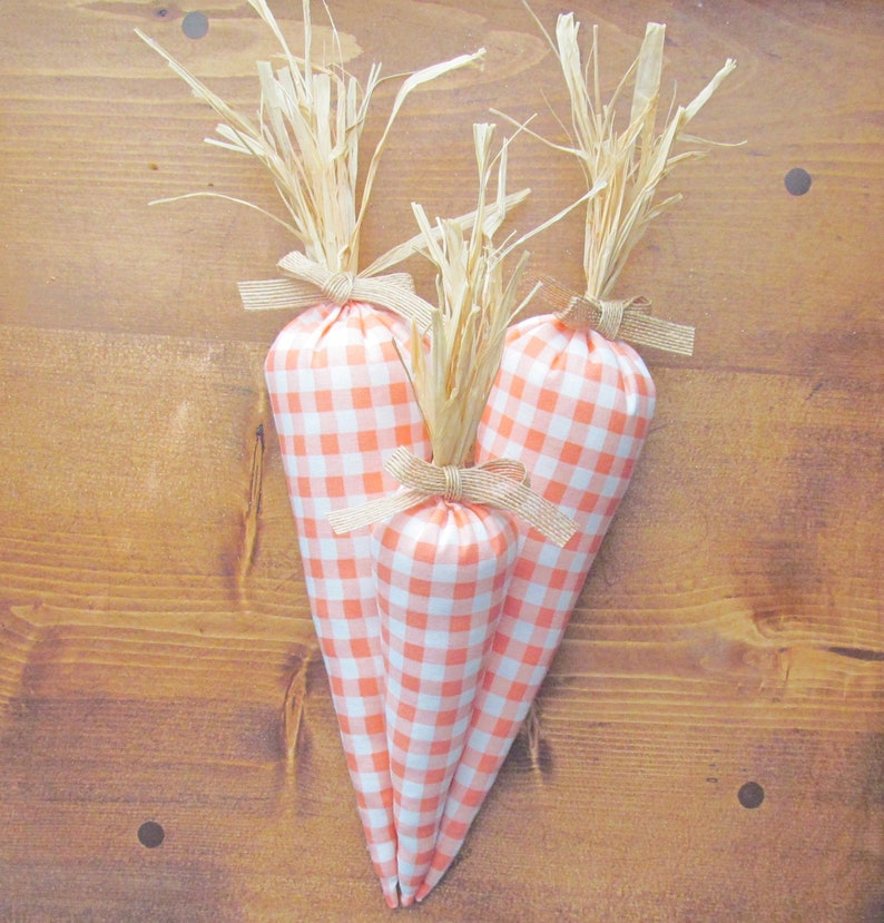 Puede incluir: Tres zanahorias de tela con un patr&oacute;n de cuadros vichy en tonos naranja y blanco. Las zanahorias est&aacute;n atadas con una cinta marr&oacute;n y tienen puntas de paja.