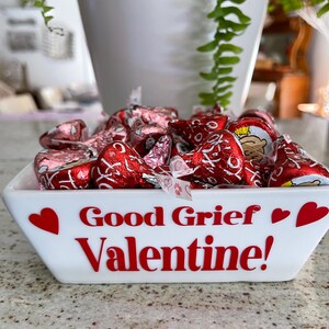 May include: White ceramic bowl with red hearts and the text "Good Grief Valentine!" filled with red and pink foil-wrapped chocolate candies.