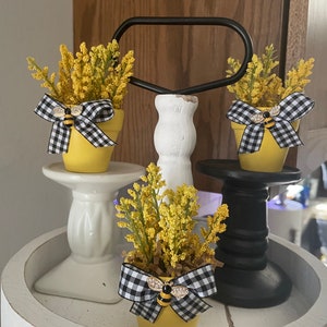 May include: Three yellow flower pots with black and white checkered bows and bee accents on a tiered stand. The stand is made of white and black wood.