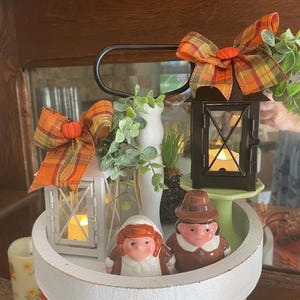 May include: Two-tiered white tray with a fall theme. The top tier features two pilgrim figurines, a small white lantern with a lit candle, a small green plant, and a black lantern with a lit candle. The bottom tier has two candles and a burlap bow.
