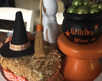May include: A tiered tray decorated for Halloween with a black witch hat, a broom, a small cauldron filled with green glitter balls, and a sign that reads "Witches Brew".