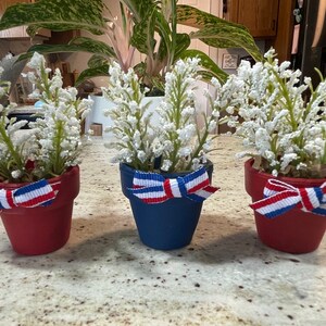 May include: Three small terracotta pots painted in red, white, and blue with white flowers and red, white, and blue ribbon bows.