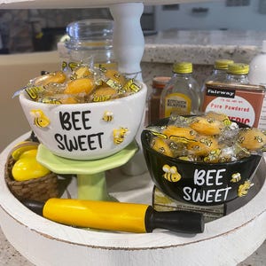 May include: Two decorative bowls with the text "BEE SWEET" and bee illustrations. One bowl is white, the other black, both filled with wrapped candies. A yellow rolling pin and other kitchen items are also visible.