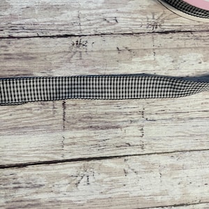 May include: Black and white gingham ribbon, a classic pattern, is displayed against a weathered, light-colored wooden surface. The ribbon is neatly rolled, showcasing its woven texture and contrasting colors. This ribbon is suitable for various crafting projects.