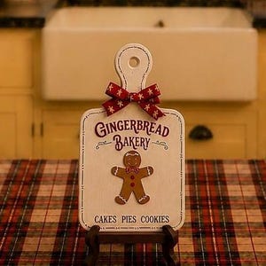 May include: A white wooden cutting board sign featuring a red ribbon bow at the top, the text GINGERBREAD BAKERY, a central gingerbread man illustration, and the text CAKES PIES COOKIES at the bottom, displayed on a small dark brown stand.