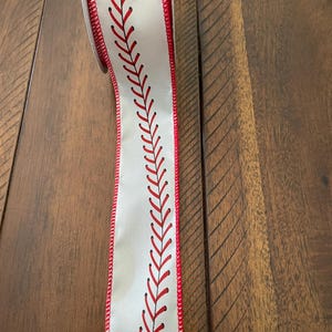 May include: A roll of white ribbon with a baseball stitch design. The ribbon has red stitching and edges, mimicking the look of a baseball. The ribbon is on a wooden surface.