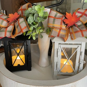 May include: Two white and black lanterns with candle lights inside, decorated with plaid bows and fall leaves on a white tiered tray.