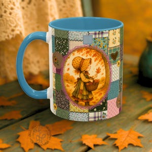May include: A ceramic mug with a blue interior and handle, featuring a patchwork design and an oval illustration of a girl in a bonnet holding a basket of leaves. The mug has a fall theme with warm colors.
