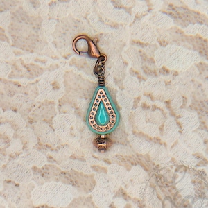 May include: A copper-colored charm with a teardrop-shaped turquoise bead. The bead is accented with small, gold-colored beads around the edges and a copper-colored bail. The charm has a lobster clasp for easy attachment.