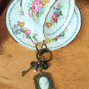 May include: A vintage-style keychain with a bronze-colored key, a cameo pendant, and a small hat charm. The keychain is paired with a floral teacup and saucer set with gold trim. The teacup and saucer have pink, yellow, and blue floral patterns.