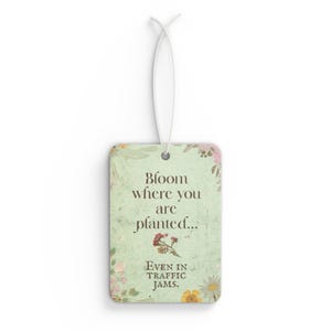 May include: Rectangular air freshener with a floral design and the text "Bloom where you are planted... Even in traffic jams." The air freshener has a white string for hanging and features a light green background.