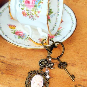May include: A vintage-style keychain with a bronze-colored frame featuring a cameo, a key, and other charms. The keychain is set against a floral teacup and saucer with gold trim. The teacup and saucer have pink, yellow, and blue floral designs.