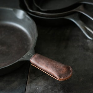 May include: A cast iron skillet with a brown leather handle. The skillet is seasoned and has a dark gray color.