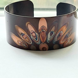Puede incluir: Un brazalete de cobre con un diseño abstracto colorido. El diseño presenta una serie de círculos concéntricos en tonos de marrón, naranja, rosa y azul.