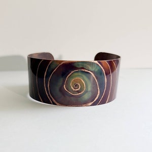 Puede incluir: Un brazalete de cobre con un diseño en espiral verde, azul y morado. El diseño está delineado en oro.