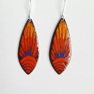 Puede incluir: Un par de pendientes en forma de lágrima con un diseño geométrico en tonos naranja, rojo y azul. Los pendientes están hechos de metal y tienen un gancho plateado.