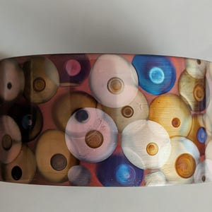 May include: A wide, cylindrical bangle bracelet featuring a translucent resin base embedded with numerous circular, bubble-like shapes in various colors including soft pink, golden yellow, deep blue, and muted brown, each with a central dot.
