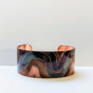 May include: A wide copper cuff bracelet with a wavy, abstract design in shades of brown, green, and copper.