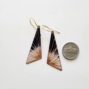 May include: A pair of gold dangle earrings with a black and white geometric design. The earrings are triangular in shape and have a white center with black lines radiating outwards. The earrings are about the size of a US quarter.