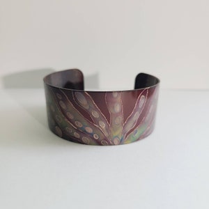Puede incluir: Un brazalete de cobre con un diseño abstracto colorido. El diseño presenta un patrón de remolinos y puntos en tonos rojos, verdes y dorados.