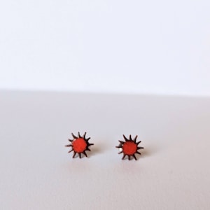 May include: A pair of small, red sun earrings with a copper finish. The earrings are in the shape of a sun with a red center and copper rays.