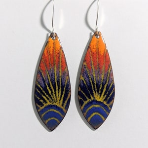 May include: A pair of teardrop-shaped earrings with a colorful sunburst design. The earrings are made of metal and have a silver hook closure. The sunburst design features shades of orange, yellow, blue, and purple.