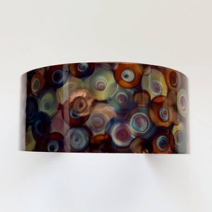 Puede incluir: Una pulsera de cobre con un patrón abstracto colorido de círculos giratorios en tonos de marrón, azul, verde y morado.