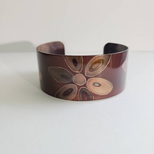 Puede incluir: Pulsera ancha de cobre con diseño floral. La pulsera presenta un fondo marrón oscuro con un patrón floral en tonos marrones, beige y cobre. El brazalete es abierto en la parte inferior y tiene un acabado liso y pulido.