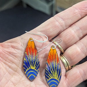 May include: A pair of teardrop-shaped earrings with a colorful enamel design. The earrings feature a blue, yellow, orange, and red sunburst pattern. The earrings are made of metal and have silver hooks.