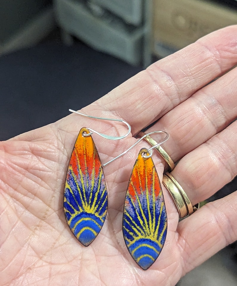 May include: A pair of teardrop-shaped earrings with a colorful enamel design. The earrings feature a blue, yellow, orange, and red sunburst pattern. The earrings are made of metal and have silver hooks.