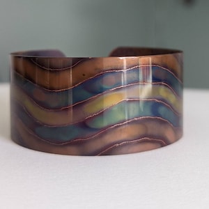 Puede incluir: Una pulsera de puño de cobre con un patrón ondulado e iridiscente en tonos marrón, azul, verde y amarillo. El metal tiene un acabado pulido y el diseño se asemeja a capas de color. Esta joya es un accesorio llamativo.