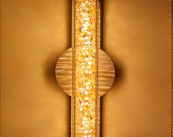 May include: A wall-mounted light fixture with a long, rectangular, clear glass shade filled with small, sparkling crystals. The fixture has a gold metal frame and a circular, wooden base.
